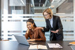 © Liubomir - Strict female boss scolding a pressured female employee looking upset and holding her head, experiencing bullying and workplace conflict in a modern office environment