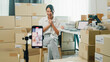 © tirachard - Youth Asia lady owner small business online shop sit front of phone look at camera live streaming talk to customer order promotion with full carton box at home. Shipping and delivery concept.
