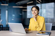 © Tetiana - Portrait of a smiling young businesswoman in a yellow shirt sitting at a desk in the office, holding her hand near her face and looking forward