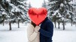 © Sophia - Romantic couple in warm winter sweaters hides their faces behind a large red heart-shaped balloon during a date in a snowy park with falling snow