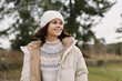 © SHOTPRIME STUDIO - Woman in a warm coat and beanie outdoors, smiling in a grassy field with trees, capturing a cozy, natural lifestyle moment in cool weather.