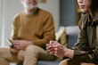 © AnnaStills - Asian young adult woman holding rosary sitting on sofa with Caucasian middle aged man in background talking, both appearing engaged in conversation during meeting indoors