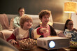 © DragonImages - Group of Caucasian and Asian children and teenagers watching movie together at home, sitting on sofa and floor, eating popcorn, smiling and interacting with projector in foreground