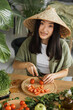 © sofiko14 - A young Asian woman wearing a conical hat is preparing fresh vegetables for a healthy meal in a bright, natural setting