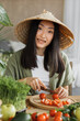 © sofiko14 - A young Asian woman wearing a conical hat is preparing fresh vegetables for a meal in a bright, natural setting