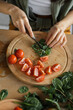 © sofiko14 - A person is carefully chopping fresh arugula and tomatoes on a wooden cutting board, preparing a healthy meal