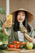 © sofiko14 - A young Asian woman wearing a conical hat drinks a green smoothie while filming herself with a smartphone surrounded by fresh ingredients