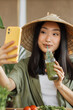 © sofiko14 - A young Asian woman wearing a conical hat drinks a green smoothie while taking a selfie with her smartphone, surrounded by fresh ingredients