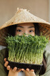 © sofiko14 - A young woman wearing a conical hat holds a tray of fresh microgreens in front of her face, showcasing healthy eating and sustainable agriculture