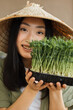 © sofiko14 - A young woman wearing a conical hat holds a tray of fresh microgreens, smiling warmly at the camera