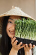 © sofiko14 - A young woman wearing a traditional conical hat smiles while holding a tray of fresh green sprouts in front of her face