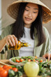 © sofiko14 - A young Asian woman wearing a conical hat adds olive oil to a fresh salad with tomatoes and greens