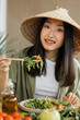 © sofiko14 - A young Asian woman wearing a conical hat enjoys a healthy salad with chopsticks, surrounded by fresh ingredients and olive oil
