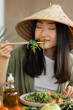 © sofiko14 - A young Asian woman wearing a conical hat enjoys a fresh green salad with cherry tomatoes and dressing, using chopsticks to pick up a bite