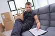 © H_Ko - In a bright living room filled with moving boxes, a focused worker sits on a gray couch, making a phone call. He reviews important documents, preparing for new beginnings.