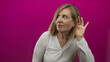 © Krakenimages.com - Middleaged woman in white top holds hand to ear in magenta studio setting; attention focus curiosity.