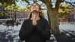 © Krakenimages.com - Young woman with hands pressed together praying on a sunlit street plaza beside a large plane tree and blurred buildings; hope gratitude faith.