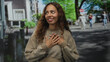 © Krakenimages.com - Young african american woman smiling with hands over chest on a city street wearing a beige sweatshirt and natural curls; gratitude warmth.