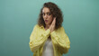 © Krakenimages.com - Young woman covering mouth with hands and wide eyes in teal studio wearing yellow shirt and tank top; surprise.