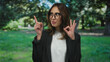 © Krakenimages.com - Hispanic woman making an ok gesture and pointing upward in a park with trees in the background, wearing glasses and a black jacket, reflecting confidence and positivity.