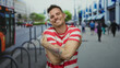 © Krakenimages.com - Young man smiling on a busy urban street with tattoos and striped shirt enjoying an outdoor city environment with people and vehicles in the background.