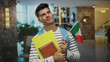 © Krakenimages.com - Young man holding an italian flag with enthusiasm inside a resort hotel, carrying documents, symbolizing travel and cultural connection in a vibrant indoor setting.