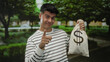© Krakenimages.com - Young man wearing striped shirt outdoors in park holding money bag while pointing enthusiastically, surrounded by lush green trees and a tranquil path in the background.