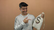 © Krakenimages.com - Young man holding money bag with dollar sign isolated against brown background smiling and showing gestures of happiness.