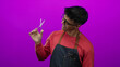 © Krakenimages.com - Young hispanic man holds hairdressing scissors against a vibrant pink background, wearing glasses, a red sweatshirt, and a denim apron, conveying modern barber style.
