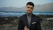 © Krakenimages.com - Young man in glasses standing on a seaside beach with mountains in the background wearing a turtleneck and blazer looking confident in a serene outdoor setting during daylight.