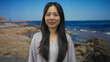 © Krakenimages.com - Young chinese woman smiling at seaside beach with clear blue sky and calm ocean waves in background showcasing tranquil outdoors setting.