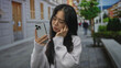 © Krakenimages.com - Young chinese woman looking at smartphone with puzzled expression on a city street outdoors, suggesting confusion or concern in urban setting.