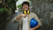 © Krakenimages.com - Smiling hispanic man pointing at a blue hardhat, wearing noise canceling headphones, stands in a picturesque mediterranean town alley.