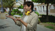 © Krakenimages.com - Hispanic man with a charming smile wearing noise-canceling headphones and extending hands on a sunny street, capturing an outdoor urban vibe with lush greenery and modern style.