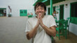 © Krakenimages.com - Hispanic man smiling on a restaurant terrace with green furniture, hands together in a joyful gesture, conveying happiness and positivity on a sunny day.