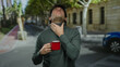 © Krakenimages.com - Hispanic man outdoors on street holding red coffee cup appearing thoughtful with trees and buildings in background.