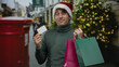 © Krakenimages.com - Hispanic man wearing a christmas hat smiling outdoors on a festive street while holding colorful shopping bags and a credit card, highlighting holiday shopping cheer.