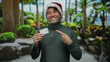 © Krakenimages.com - Hispanic man wearing a christmas hat joyfully points to a card outdoors on a lush street.