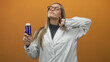 © Krakenimages.com - Woman in white lab coat holding blue medicine bottle with hand behind neck and wearing glasses against orange studio backdrop; confidence.