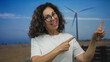 © Krakenimages.com - Woman smiling in a field with a windmill, wearing glasses and pointing, representing renewable energy and outdoor lifestyle.