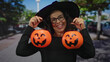 © Krakenimages.com - Woman dressed as a witch holding two pumpkin baskets on a street with trees in the background, showcasing a festive halloween scene outdoors.