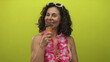 © Krakenimages.com - Hispanic woman in bikini and hawaiian lei enjoys ice cream against an isolated yellow background wall, exuding a joyful summer vibe.