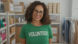 © Krakenimages.com - Woman in green volunteer shirt smiling indoors at a charity room, surrounded by organized supplies on shelves, wearing glasses and exuding warmth.