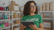 © Krakenimages.com - Woman volunteer wearing green uniform smiles while using tablet in charity room with shelves full of donations like food and supplies in an indoor setting.