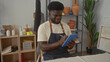 © Krakenimages.com - Man in denim apron writes on blue tablet with stylus in studio among pottery shelves and plants; creative concentration.