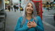© Krakenimages.com - Senior woman with grey hair standing on a busy london street in front of a classic red phone booth, expressing emotion with her hands clasped to her chest.