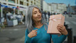 © Krakenimages.com - Woman with grey hair points at pink gift box while standing on a busy street, highlighting her joyful expression and the vibrant urban environment around her.