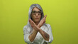© Krakenimages.com - Senior woman with grey hair and tattooed arm poses with crossed arms against vibrant yellow background, wearing glasses and expressing disapproval or defiance with confidence.