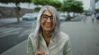 © Krakenimages.com - Senior woman with grey hair smiling outdoors on a city street with cars and trees in the background, showcasing a joyful and casual urban scene.