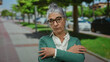 © Krakenimages.com - Senior woman with grey hair stands outdoors on a city street, arms crossed, wearing glasses and a green cardigan, embodying confidence amidst urban greenery and parked cars.